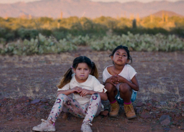 Two young girls sit in dirt and rocks, with grasses and mountains visible in the background. 