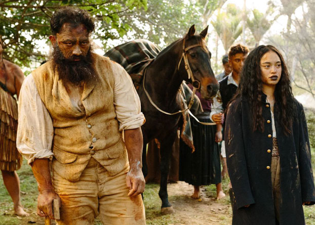 A man with a long beard and a woman with black lips stand. A brown horse stands behind them. 