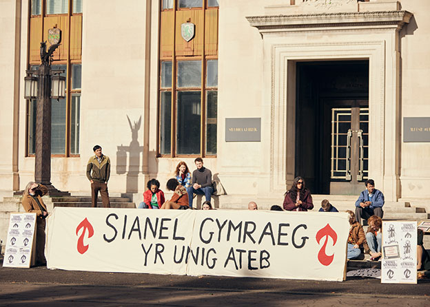 A group of people sit outside on the steps of a white building behind a large sign in Welsh.