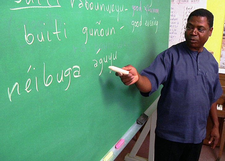 A man points to a green chalkboard with a piece of chalk. On it are written foreign words and their English translations.