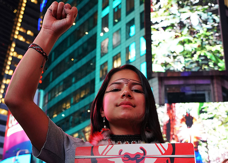 A young woman with tribal markings across her forehead raises her fist, holding up a sign on a city street.