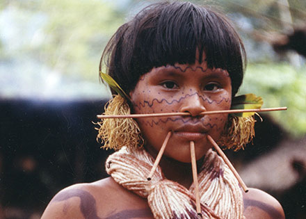Young person with patterned tattoos across their face and chest, with wooden spear-like piercing through their septum and lower lip.