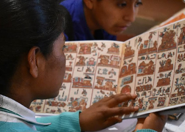A young person holds a book and studies the colorful drawings and designs on the page intently. 