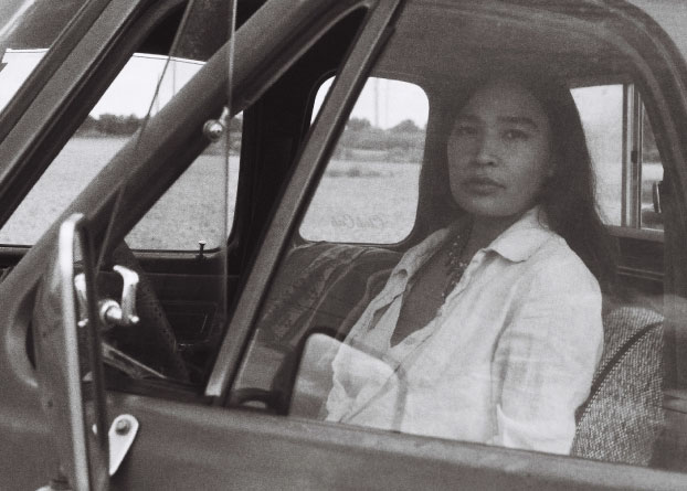 A woman looks directly into the camera while sitting behind the wheel of a car. 