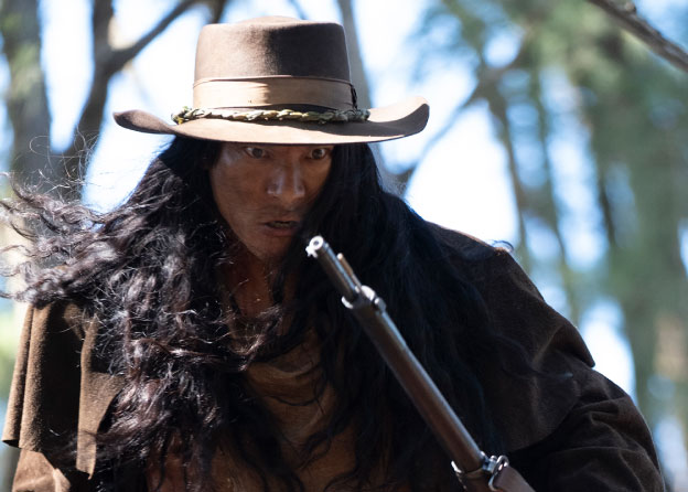 A person with long dark hair wears a hat and holds a rifle. His expression indicates anger, and he appears to be in motion.