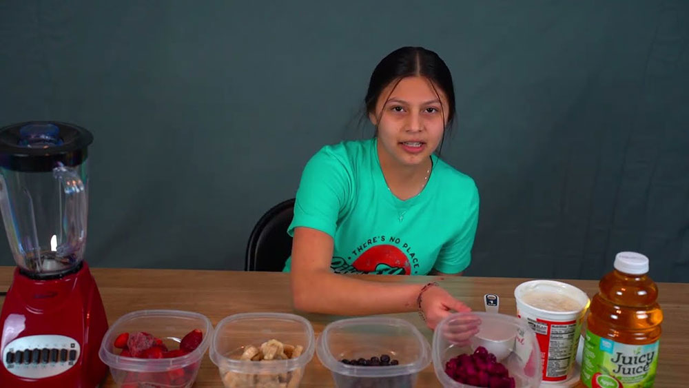 A young girl sits at a table with containers of berries, juice, and a blender.