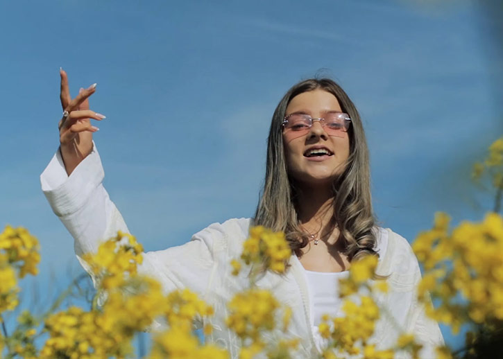 A woman in a long-sleeved white top and pink shades, with long dark hair, sings among yellow wildflowers.